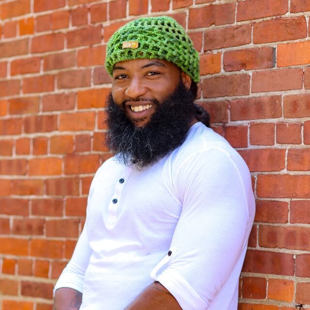Deonte Cooley smiling in a green knit cap and white shirt, leaning against a brick wall